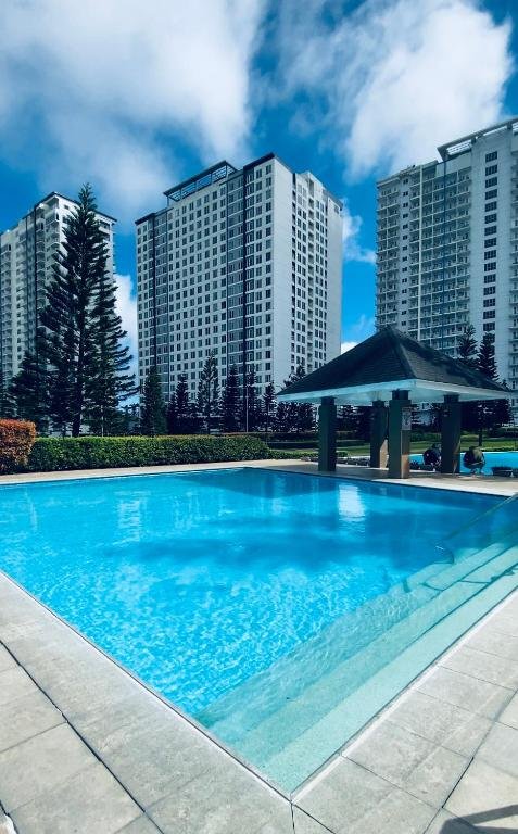Outdoor swimming pool at Tagaytay Staycation by SMDC Wind Residences Lakeview in the Philippines, featuring a covered poolside gazebo, pine trees, and modern high-rise condo towers under a cloudy blue sky.