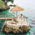 Vibrant daytime landscape photograph capturing the coastal access area at Stay N Save B&B in Cebu, Philippines. A person in a white top and dark shorts sits relaxing on a concrete viewing platform with a log railing, built onto a natural rock formation in crystal-clear turquoise and deep blue ocean waters. The platform features a large Nipa palm thatch umbrella and a slatted bamboo lounge chair. A rustic, weathered bamboo plank footbridge spans from the lush green cliffside over the emerald water to the platform. Rocky cliffs and coastal vegetation frame the scene under a bright sky.