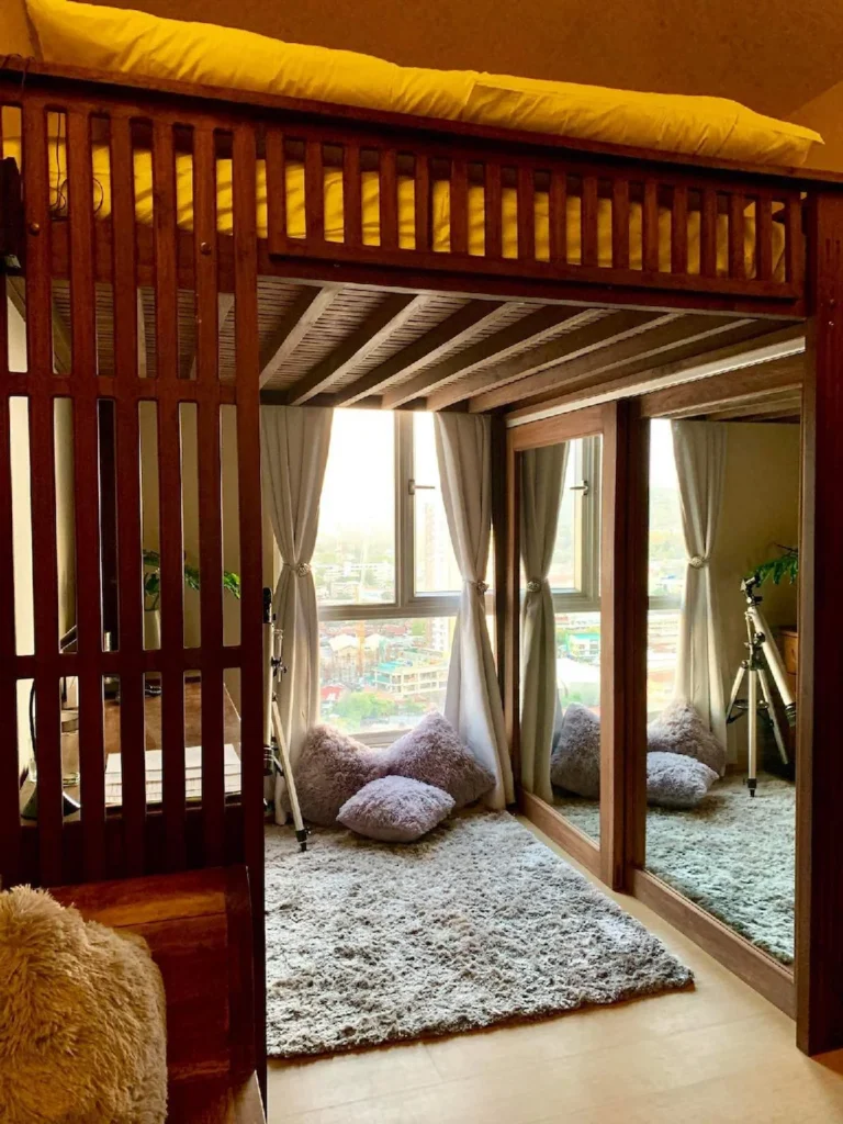 A view into a compact studio penthouse condo at Centrio Tower in Cagayan de Oro, Philippines, featuring a large, dark wood loft bed structure. Below the bed is a cozy nook with a grey shag rug, three fluffy purple pillows, a window with curtains and a city view, and a small telescope. To the right is a wall of floor-to-ceiling mirrored closet doors.