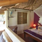 Interior view of a rustic guest room at Sugar Lounge in Sipalay, Philippines, featuring a bed with purple accents and a mosquito net, a bamboo ceiling, and an open-air seating area overlooking a tropical garden.