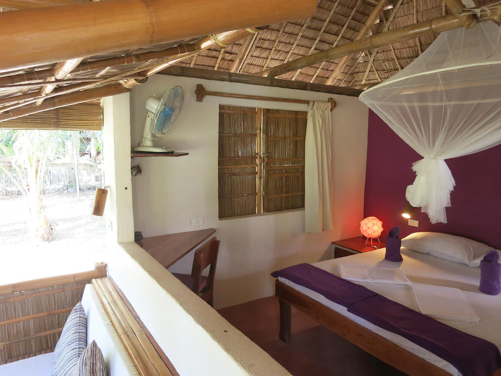 Interior view of a rustic guest room at Sugar Lounge in Sipalay, Philippines, featuring a bed with purple accents and a mosquito net, a bamboo ceiling, and an open-air seating area overlooking a tropical garden.
