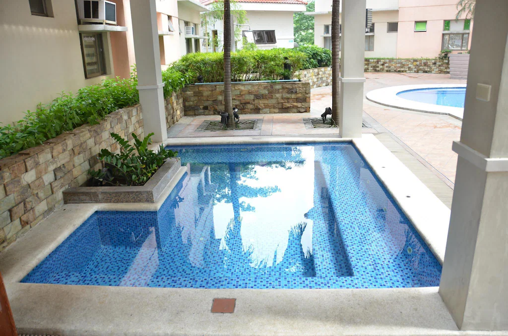 Outdoor swimming pool at A Homey Place Manila hotel, surrounded by lush tropical plants and lounge chairs under a clear blue sky in Manila, Philippines.