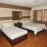 A hotel room interior at The Alpa Hotel and Restaurant in Batangas, Philippines, featuring one single bed and one double bed with white linens, dark wood furniture, and colorful patterned curtains.