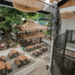 High-angle view of an open-air hostel courtyard featuring wooden picnic tables, a thatched-roof native pavilion, and a spiral staircase