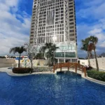 Exterior view of The Excelsior Hotel in Las Pinas, Metro Manila, featuring a modern high-rise building and a beautifully designed swimming pool with a wooden bridge and palm trees under a sunny sky.