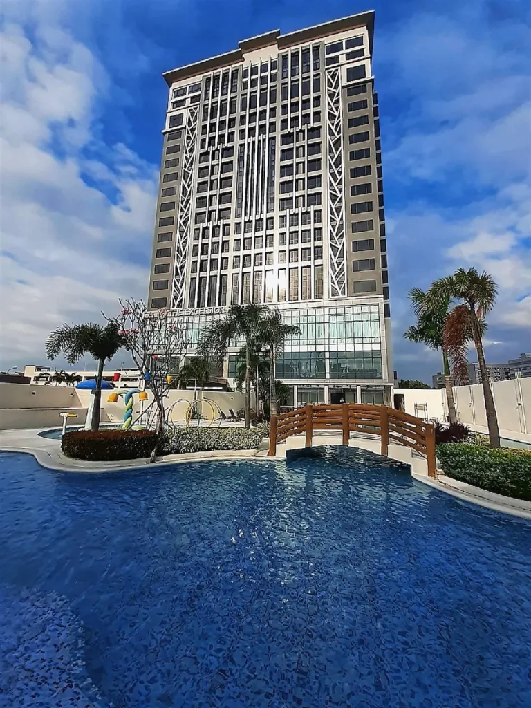 Exterior view of The Excelsior Hotel in Las Pinas, Metro Manila, featuring a modern high-rise building and a beautifully designed swimming pool with a wooden bridge and palm trees under a sunny sky.