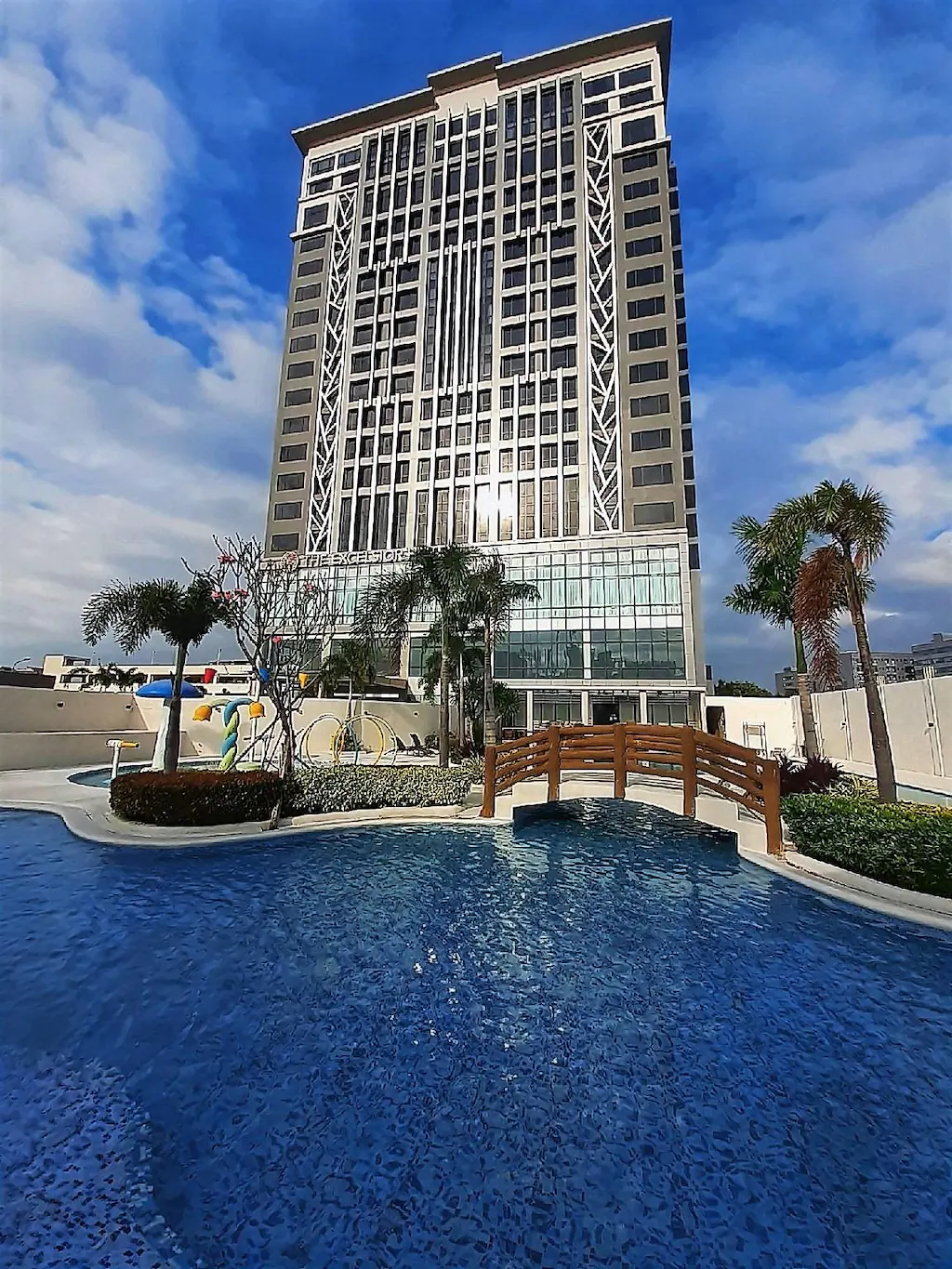 Exterior view of The Excelsior Hotel in Las Pinas, Metro Manila, featuring a modern high-rise building and a beautifully designed swimming pool with a wooden bridge and palm trees under a sunny sky.