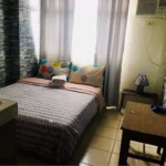 Compact and cozy studio apartment interior at Urban Deca Tower, Manila, Philippines, showing a made bed with graphic pillows and geometric duvet, a small wooden table, a wall-mounted TV, window, and a kitchen sink area, all on tiled floors with stone-patterned wallpaper.