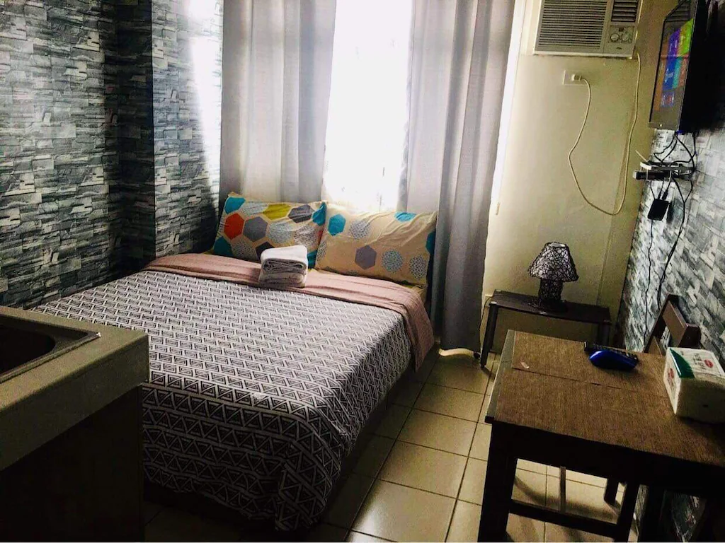 Compact and cozy studio apartment interior at Urban Deca Tower, Manila, Philippines, showing a made bed with graphic pillows and geometric duvet, a small wooden table, a wall-mounted TV, window, and a kitchen sink area, all on tiled floors with stone-patterned wallpaper.