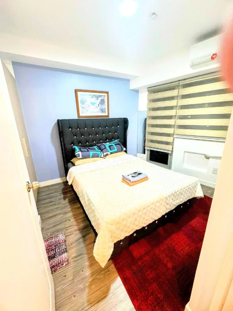 Modern and cozy bedroom at Urban Oasis in The Residences Commonwealth, Quezon City, Manila, featuring a dressed queen bed, warm ambient lighting, large windows with city views, and elegant minimalist decor.