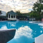 The main swimming pool at V Resort Dasma in Cavite, Philippines, perfectly reflecting a dramatic sunset sky with clouds, surrounded by lush palm trees, a gazebo with a viewpoint, and vibrant colorful planters, promising a relaxing vacation experience.