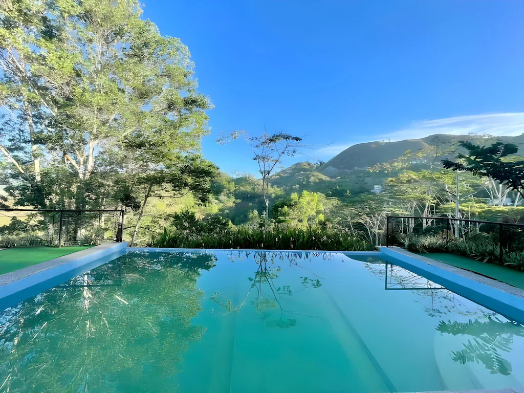 An outdoor infinity swimming pool reflecting a vibrant blue sky and green trees with mountains in the background at Victoria's Peak - El Cinco Farmville and Resort.