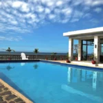 A large blue outdoor swimming pool at Villas Buenavista in La Union, overlooking the sea with white lounge chairs along a stone deck under a cloudy sky.