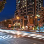 A nighttime photograph of the Vivere Hotel, Manila, Philippines, showing its modern multi-story building, curved gold entrance structure, and palm-tree-lined avenue with long light streaks from moving vehicles.