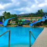 A vibrant outdoor swimming pool area at YesGo Cold Spring Resort in Maragusan, Philippines, featuring clear blue spring water, stainless steel steps, two bright blue water slides, and a colorful terraced backdrop with resort signage.