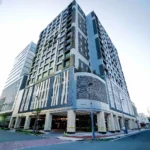 Modern high-rise facade of Bayprime Hotel Manila, featuring contemporary architecture with white, gray, and dark accents, an open street corner view, and street lamps.