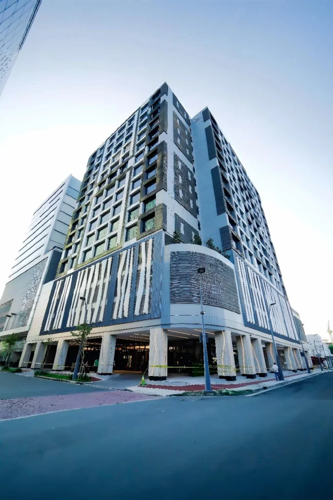 Modern high-rise facade of Bayprime Hotel Manila, featuring contemporary architecture with white, gray, and dark accents, an open street corner view, and street lamps.