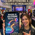 A smiling concertgoer in a black leather jacket shows a smartphone map indicating Bayprime Hotel is a 5-minute walk from the SM Mall of Asia Arena lobby.