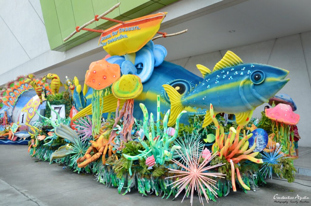 A vibrant, ocean-themed parade float featuring a large blue and yellow tuna fish sculpture, colorful coral reefs, and a traditional outrigger boat during the Tuna Festival.