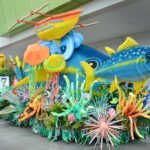 A vibrant, ocean-themed parade float featuring a large blue and yellow tuna fish sculpture, colorful coral reefs, and a traditional outrigger boat during the Tuna Festival.